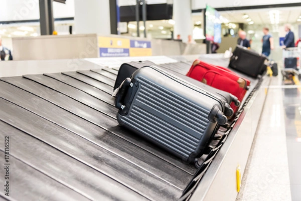 Fototapeta Suitcase on luggage conveyor belt at baggage claim at airport. Lines of people waiting for their baggage