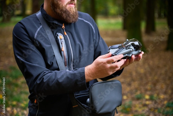 Fototapeta A man in a black jacket examines his drone carefully, surrounded by vibrant trees and fallen leaves in a serene park setting.