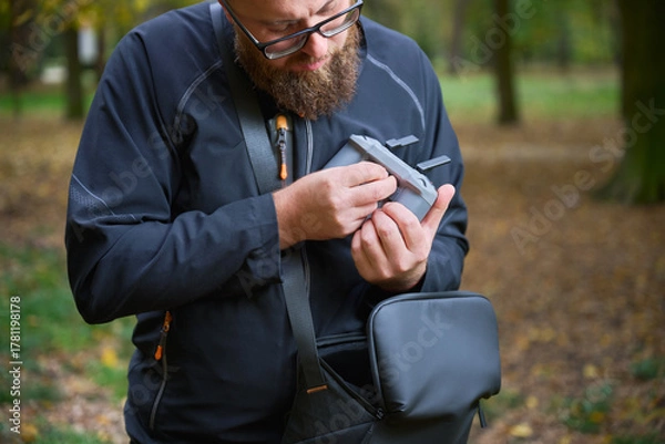 Fototapeta A person examines a camera accessory closely while standing in a beautiful autumn park. Surrounding yellow and orange leaves create a peaceful atmosphere.