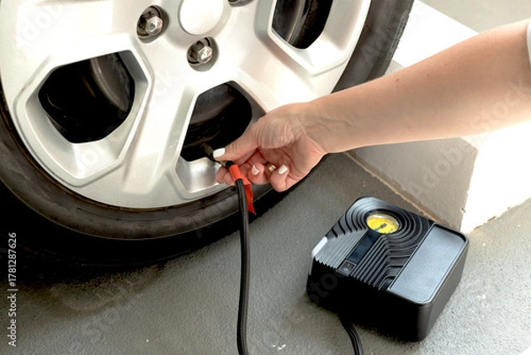 Fototapeta Close-up of woman inflating car tire using portable electric air compressor. DIY vehicle maintenance at home or garage.