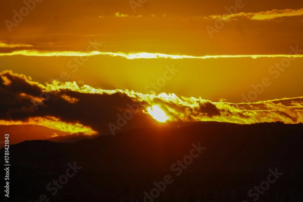 Obraz A powerful sunset paints the sky in glowing gold and deep orange hues over the dark silhouettes of hills.
