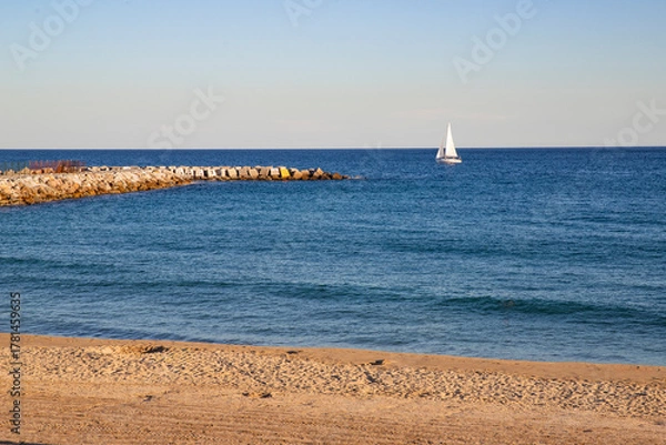 Obraz A tranquil seaside landscape with a sandy beach and a stone breakwater. A serene landscape with a yacht sailing on a calm sea