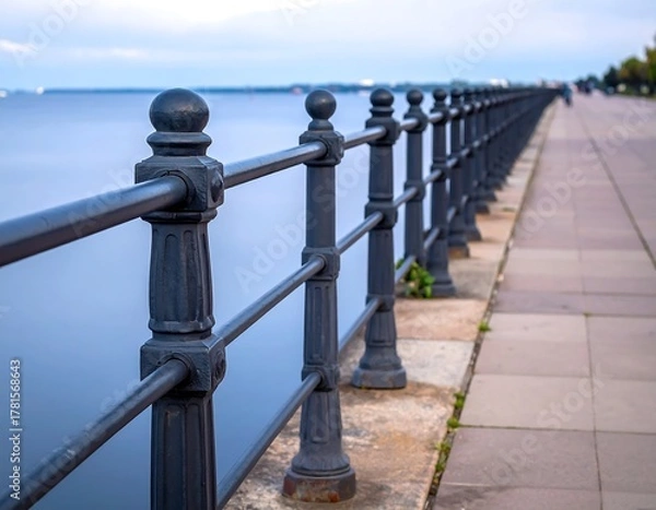 Fototapeta Serene waterfront scene with a black railing, blurred water, walkway, & sky