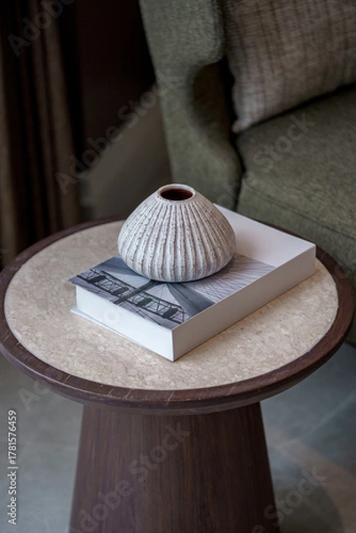 Fototapeta Cozy living room decor. A textured ceramic vase sits on a book, placed on a round wood and marble side table. Blurred armchair in background.