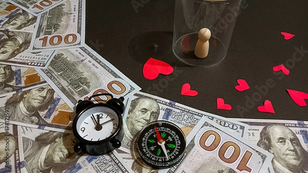 Fototapeta A creative composition featuring a small clock, compass, dollar bills, red paper hearts, and a wooden figure under a glass, symbolizing the connection between time, love, and financial direction.