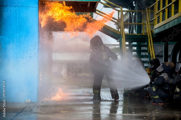 Fototapeta Firemen using water from hose for fire fighting at firefight training of insurance group. Firefighter wearing a fire suit for safety under the danger case.