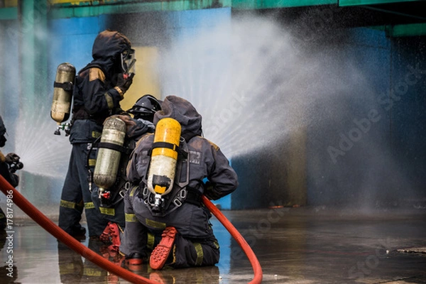 Fototapeta Firemen using water from hose for fire fighting at firefight training of insurance group. Firefighter wearing a fire suit for safety under the danger case.