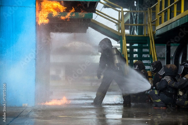 Fototapeta Firemen using water from hose for fire fighting at firefight training of insurance group. Firefighter wearing a fire suit for safety under the danger case.