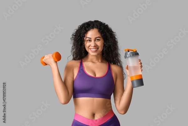 Fototapeta Sporty young African-American female coach with dumbbell and water bottle on white background