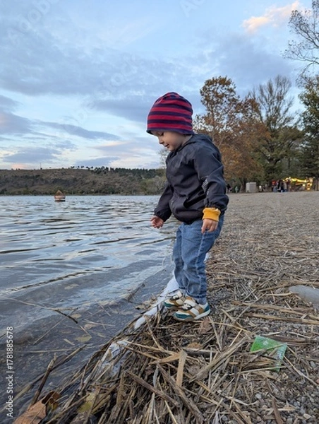 Fototapeta A two-year-old boy playing peacefully on the pebbled shore of a clear mountain lake, autumn sunshine.

