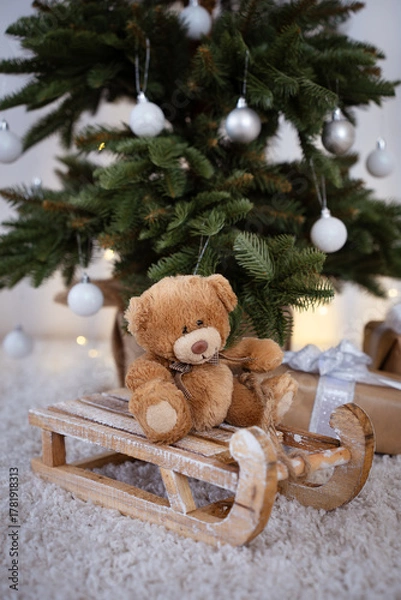 Obraz Brown vintage Teddy bear sitting on wooden sledge with Christmas tree and Christmas lights and presents in the background.	

