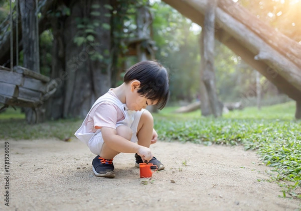 Fototapeta little boy playing in the sand with toy trucks and a shovel under dappled sunlight, enjoying a peaceful outdoor moment surrounded by trees and nature 