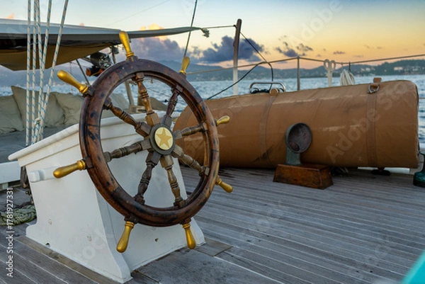 Obraz An unmanned wooden yacht wheel with gold handles glows in the Caribbean sunset, the island beyond bathed in warm light and tranquillity