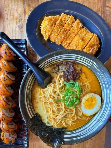 Obraz Japanese ramen with rich broth, soft-boiled egg, and seaweed served alongside crispy katsu and spicy sushi rolls. A vibrant Asian meal representing flavor, comfort, and culinary craftsmanship.