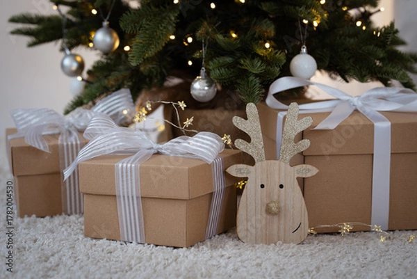 Obraz A wooden reindeer decoration with gold glitter details and Christmas present with white ribbon under a Christmas tree with lights in the background. Christmas decorations. Christmas theme.