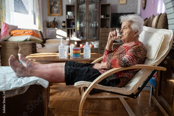 Fototapeta This image captures a senior woman relaxing in an armchair with her feet up, thoughtfully using an inhaler, highlighting the importance of respiratory health and familial care indoors.