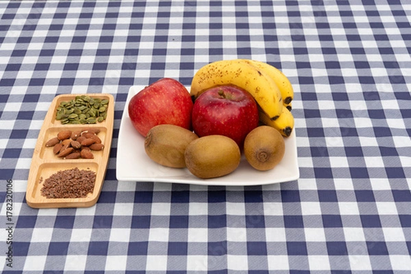 Obraz Platter of fruit (apples, kiwis, bananas) and a divided tray with almonds, pumpkin seeds, and flax seeds on a navy blue and white plaid tablecloth. Ideal for healthy snack and diet concepts.