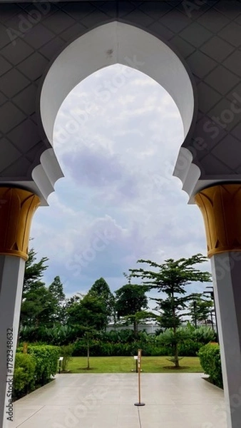 Obraz Scenic view through a mosque arch revealing a green garden and cloudy sky. A peaceful composition uniting Islamic architecture and nature’s calm beauty.
