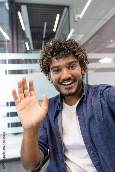 Fototapeta Vertical close-up photo of a young Indian man in an office talking to a camera he is holding in his hand