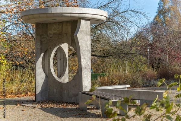 Fototapeta Modern concrete pavilion with circular cutouts and benches in a park. Late-autumn shrubs and bare branches surround it.