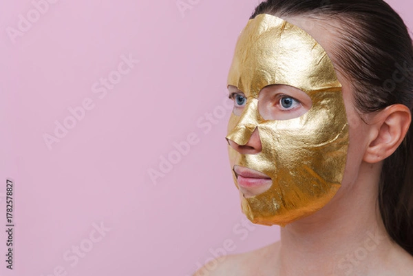 Fototapeta A woman with a golden facial mask against a pink background. The image represents self‑care, beauty and personal wellness. Copy space