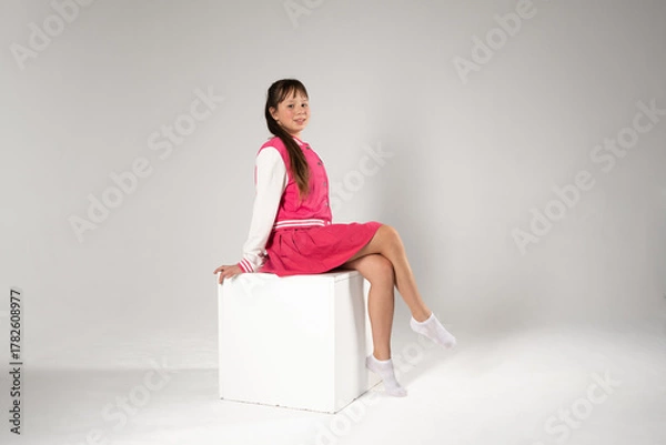 Fototapeta A full-length studio portrait of a smiling young girl with long brown hair, wearing a pink and white varsity jacket, a matching pink skirt, and white socks, sitting elegantly on a white cube against a