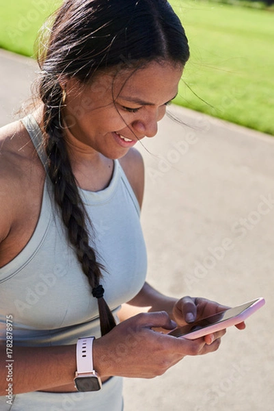 Obraz Smiling african millennial woman using smartphone in a park after training, wearing sportswear and smartwatch. Close up side view conveys mobile phone connection, positivity healthy active lifestyle.