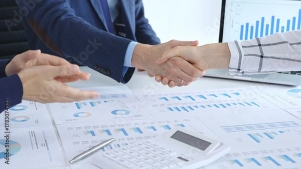 Fototapeta Business people shaking hands over financial documents and laptop, confirming successful partnership, agreement, or merger in a corporate office meeting
