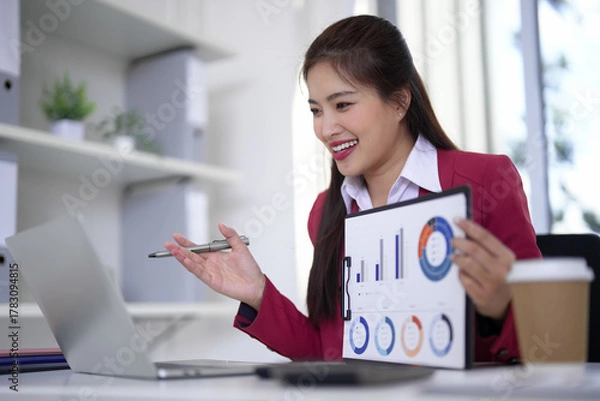 Fototapeta Asian business professional presents financial data online meeting uses laptop computer modern office setting successful women analyzes report performance