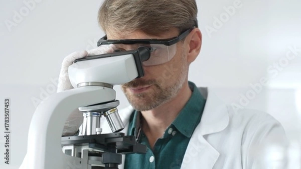 Fototapeta Senior man scientist wearing protective glasses and lab coat analyzing samples with a microscope in laboratory setting. Medicine, health care and science concept