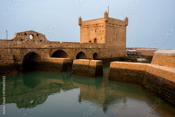 Fototapeta Wide golden hour shot of the stone bridge leading to the Castelo Real and the calm inner harbor of Essaouira, Morocco. The warm sunlight enhances the textures of the fort and water reflections.