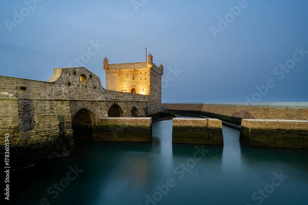 Fototapeta Wide evening long exposure shot of the stone bridge leading to the illuminated Castelo Real and the calm inner harbor of Essaouira, Morocco. The reflections of the lights shimmer across the harbor
