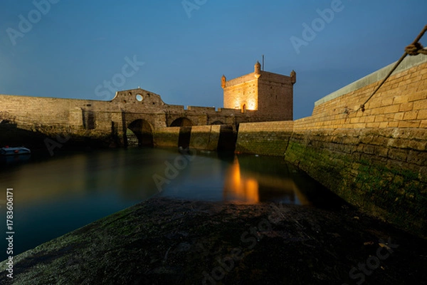 Fototapeta Long exposure night shot of the illuminated Castelo Real and the old harbor of Essaouira, Morocco. The glowing lights reflect softly on the calm water, creating a serene coastal night scene.