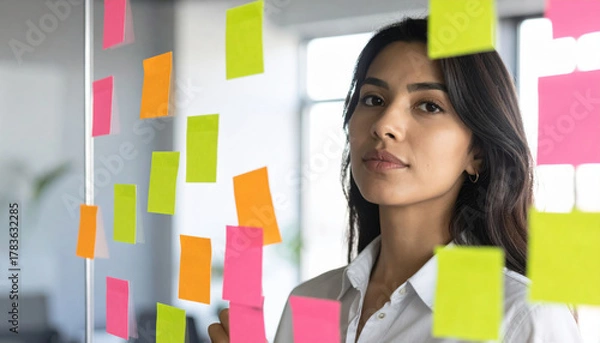 Fototapeta Focused woman stands in front of glass wall covered with colorful sticky notes, planning and organizing ideas in modern office setting