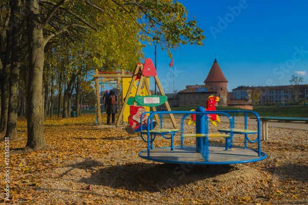 Obraz Grandmother with a grandson playing on a children playground consisting of rotating carousel, swing, tube and climbing frame. With Fallen autumn leaves of trees on the background