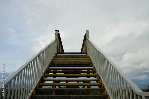 Fototapeta Weathered Outdoor Staircase With White Railings And Rusty Metal Leading Upwards To A Cloudy Sky In Towyn, North Wales, Uk