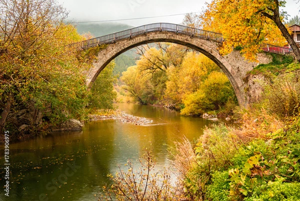Fototapeta Iconic Single-Arch Stone Bridge of Vovousa Village Spanning the Aoos River, Surrounded by Golden Autumn Foliage in Pindos, Greece