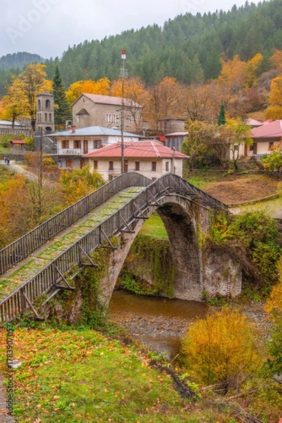 Fototapeta Vertical View of the Historic Stone Bridge and Traditional Houses of Vovousa Village, Pindos, Greece, During a Colorful Autumn Day