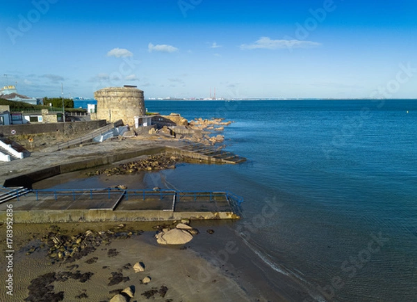 Fototapeta A beach with a dock and Martello Tower Number 14, Seapoint, Blackrock, Dublin. The water is calm and the sky is clear.