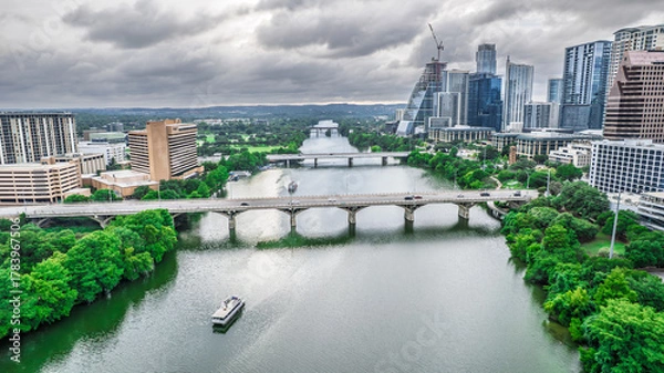 Fototapeta Panoramic aerial shot of Austin, Texas. The Colorado River flows beneath a bridge with city skyscrapers in the background under dramatic cloudy skies. Modern urban landscape, river views, Texas.