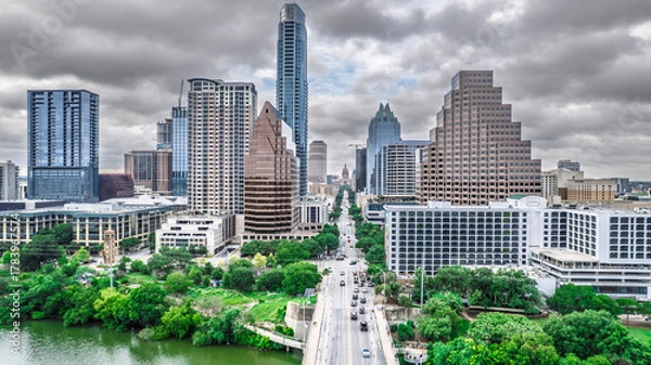 Obraz Panoramic aerial view of Austin, Texas skyline under dramatic cloudy skies. Skyscrapers dominate the cityscape alongside a busy street and lush green riverbanks. A vibrant urban scene showcasing
