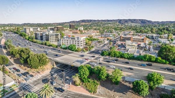 Fototapeta Aerial view of a busy highway cutting through a suburban landscape with buildings and hills in the distance. Cars flow along the road under an overpass on a sunny day. Ideal for business
