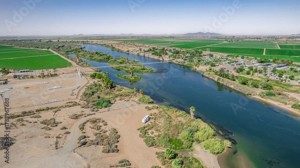 Fototapeta Aerial view of a river landscape in Arizona. Agricultural fields stretch across the horizon under a clear blue sky. A bridge spans the waterway with a small RV visible. Ideal for nature, agriculture
