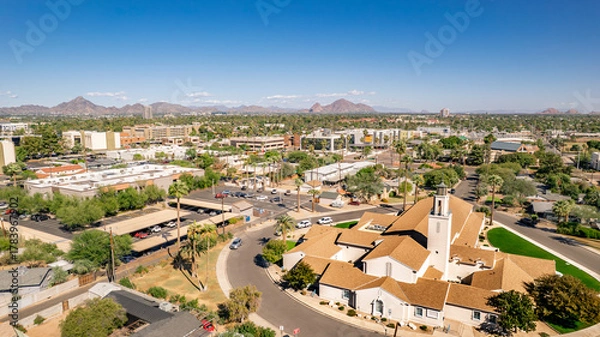 Fototapeta Aerial view of a suburban cityscape in Phoenix, Arizona. A church stands prominently amidst residential streets and palm trees under a clear blue sky. Rolling mountains form a distant backdrop