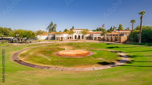 Fototapeta Panoramic view of a historic building in a desert park. A circular paved area leads to the stately structure under a clear blue sky. Palm trees and lush greenery complete this serene scene. Perfect