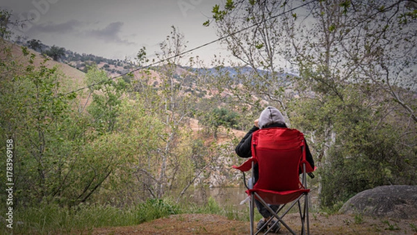 Fototapeta A lone backpacker sits in a folding chair overlooking a serene river valley. Moody landscape, reflection, contemplation, nature, peaceful moment.
