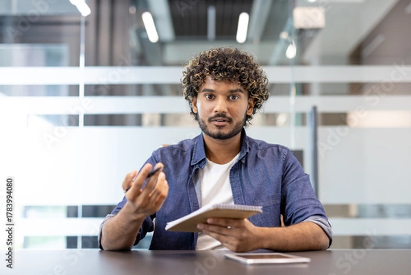 Fototapeta Portrait of a young Indian man sitting in an office in front of the camera, talking online and taking notes in a notebook