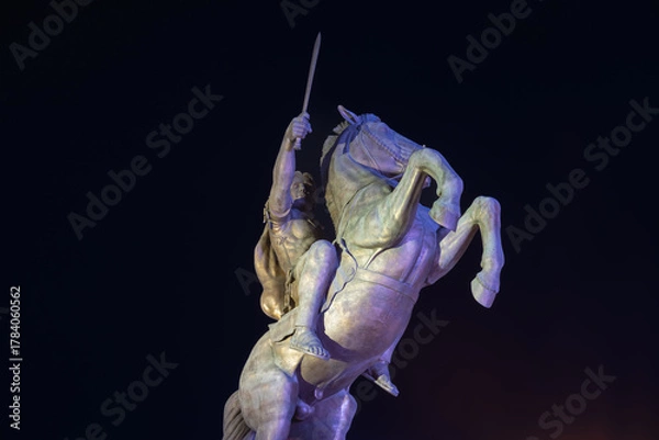 Fototapeta young warrior on a rearing horse, holding a sword high, dramatically lit at night against a dark sky, a powerful symbol of heroism and ancient history