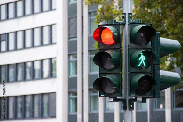 Obraz Traffic light displaying red and green signals for vehicles and pedestrians, positioned on a street corner, with modern buildings in the background, illustrating urban traffic management