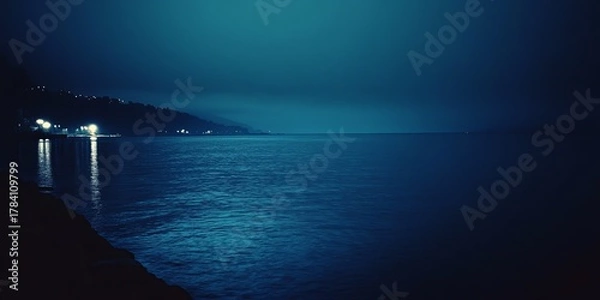 Obraz Nighttime coastal scene with hazy, dark blue water and illuminated shoreline